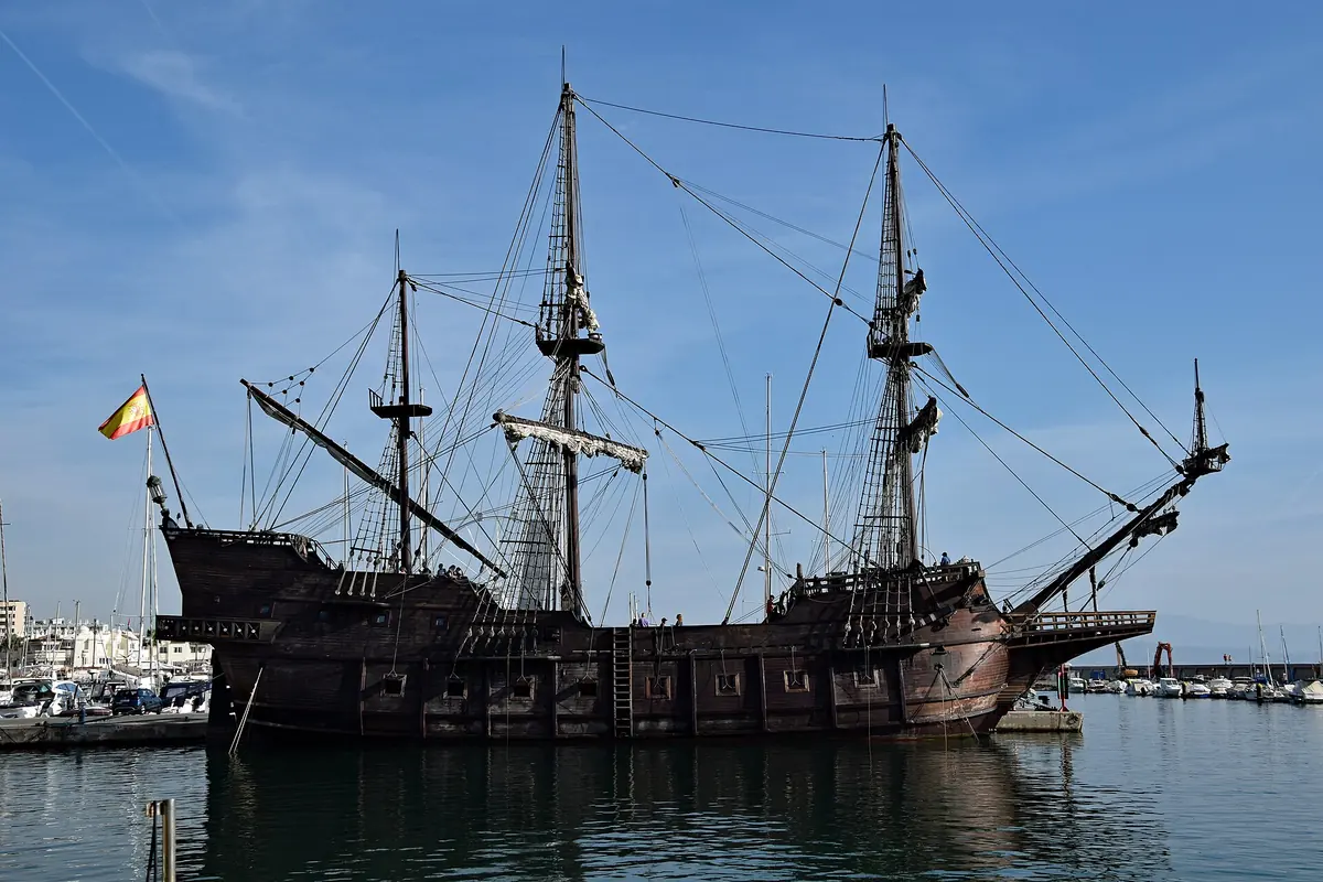 Eine Nachbau-Galeone mit drei hohen Masten und einer spanischen Flagge am Heck, vertäut in einem ruhigen Hafen